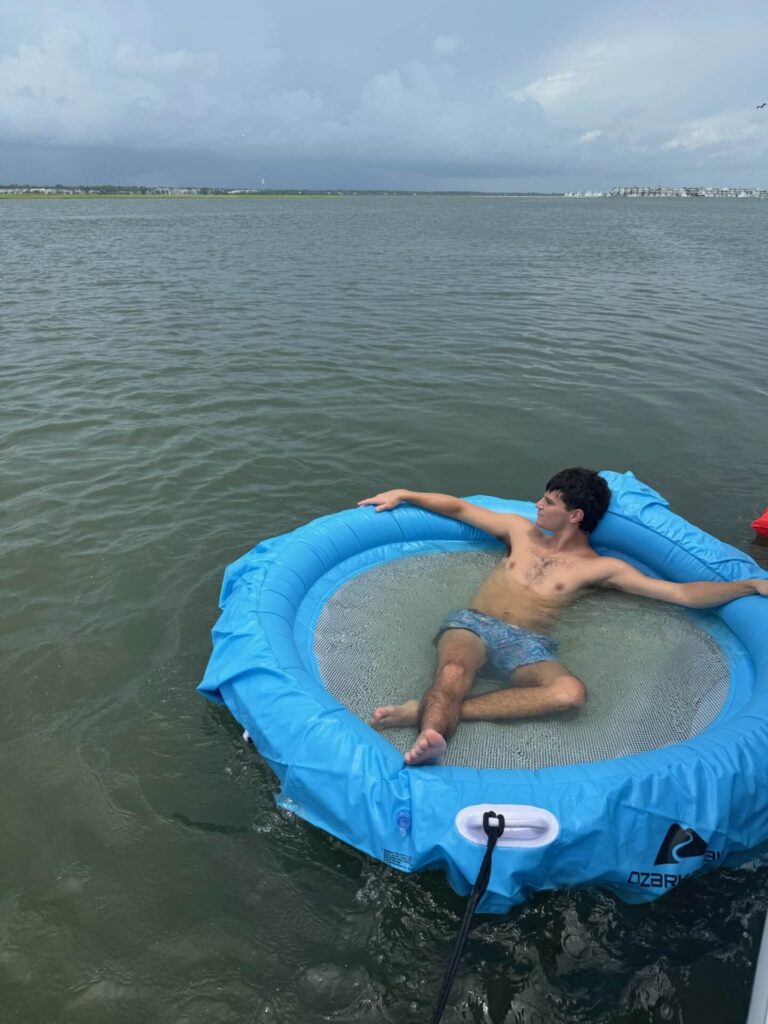 A man reclines on a blue floating water trampoline in a bay under a cloudy sky.